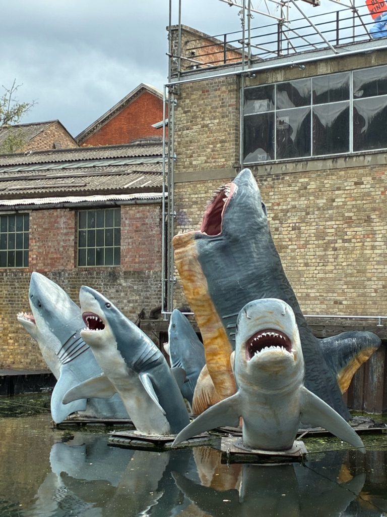 Shark sculptures emerging from canal water in front of a brick warehouse
